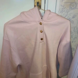 Ljusrosa hoodie från Amisu med knappar - Säljer en ljusrosa hoodie från Amisu i storlek S. Tröjan har huva, tre guldiga knappar framtill och en stor magficka. Materialet är mjukt och känns som bomull, perfekt för chill dagar. Passformen är normal och ärmarna är långa.