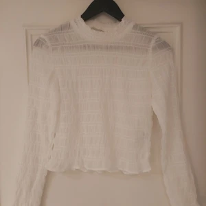 Vit blus från H&M med struktur - Säljer en vit blus från H&M i storlek M. Blusen har ett transparent, strukturerat tyg med smockdetaljer och lång ärm. Den har en rundad hals och är lätt croppad i modellen. Perfekt för dig som gillar en clean och fräsch look.