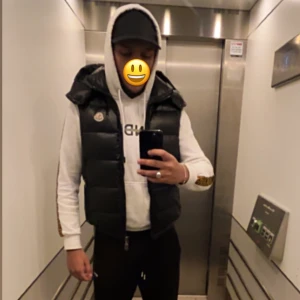 Moncler väst - Säljer en svart, glansig dunväst från Moncler med klassisk logga på bröstet. Västen har hög krage, dragkedja framtill och en quiltad design som ger en cool och sportig look. Perfekt att slänga över en hoodie för extra stilpoäng.