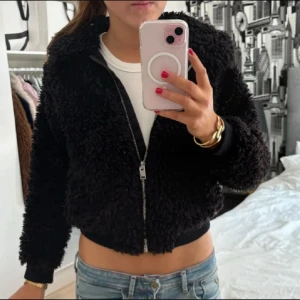 Svart teddyjacka med dragkedja - Mysig svart teddyjacka med fluffig struktur och dragkedja framtill. Jackan har en croppad passform och långa ärmar, perfekt för att hålla värmen och ändå se cool ut. Passar till jeans och sneakers för en avslappnad look.