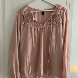 Puderrosa blus med spetsdetaljer - Supersöt puderrosa blus  med v-ringning och knytband framtill. Blusen har långa ärmar och vackra virkade spetsdetaljer över bröstet. Perfekt när man vill vara lite mer uppklädd. Tunnt material. 
