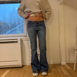 Blå bootcut jeans från Vigoss - Snygga blå jeans från Vigoss med låg midja och bootcut-ben. Klassisk femficksdesign med coola detaljer på fickorna och slitningar för en avslappnad vibe. Jeansen är gjorda i jeansmaterial med kontrastsömmar och metallknappar. Följande är jeansens mått: ytterbensmått: 101 cm innerbensmått:  80 cm Midjemått tvärs över: 35 cm Lårmått: 19 cm Grenmått: 20 cm (Nr 31)