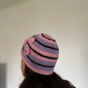 Randig virkad mössa med blommor - Supergullig virkad mössa med rosa och lila ränder, detaljerad med svarta linjer och tre virkade blommor på sidan. Perfekt för dig som vill sticka ut med en färgglad och unik accessoar. Mjuk och skön, passar till både chill och kreativ stil.
