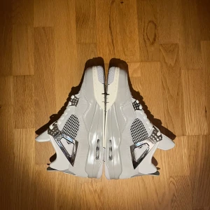 Nike Air Jordan 4 ”Retro Frozen Moments” - Snygga Nike Air Jordan 4 i vitt och silver med coola metallic-detaljer. Skorna har meshpaneler, snörning och en chunky sula. Ikonisk Jumpman-logga på baksidan och tungan. Perfekta för dig som vill sticka ut med stilrena och moderna sneakers.