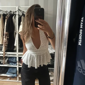 Vit prickig halterneck peplumtop - Supersöt vit halterneck peplumtop med svarta prickar och djup v-ringning. Toppen är ärmlös och har öppen rygg, vilket ger en luftig känsla. Perfekt till jeans eller kjol för en fräsch sommarlook.