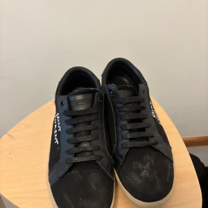 Svarta Saint Laurent sneakers - Svarta sneakers från Saint Laurent med vit logga broderad på sidan. Skorna har svart snörning, rund tå och beige platt sula. Ovandelen är i en mix av skinn och textil för en clean och stilren look. Perfekta för dig som gillar exklusiva streetwear-vibbar.