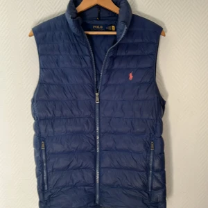 Blå dunväst från Polo Ralph Lauren - Snygg marinblå dunväst från Polo Ralph Lauren med quiltad design och dragkedja framtill. Västen har två fickor med dragkedja och klassisk logga i rött på bröstet. Perfekt för lager-på-lager och stilren look.