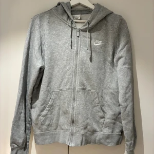 Grå hoodie från Nike med dragkedja - Grå hoodie från Nike med broderad logga på bröstet, huva med snörning och praktiska fickor framtill. Tröjan har dragkedja hela vägen och är långärmad. Perfekt för chill eller träning, klassisk stil och mjukt material. Tröjan har en fläck på vänstra axeln, samt även vid ena ärmens slut. Har därför sänkt priset. Annars mycket gott skick! 