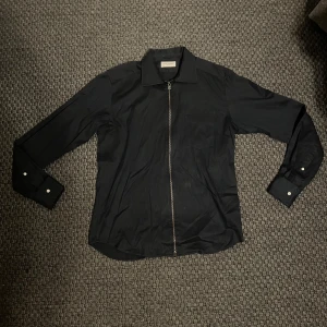 Svart skjorta med dragkedja John Henric - Svart skjorta från John Henric med klassisk krage och hel dragkedja framtill. Skjortan har långa ärmar med manschett och är tillverkad i ett slätt tyg som känns stilrent och modernt. Perfekt för dig som vill ha en clean look med edge.