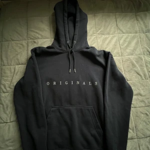 Svart hoodie från Jack & Jones Originals - Mörk blå hoodie från Jack & Jones Originals med vit text på bröstet. Klassisk modell med huva, snörning och stor magficka framtill. Mjuk insida och långärmad design, perfekt för chill dagar eller när du vill ha en clean look.