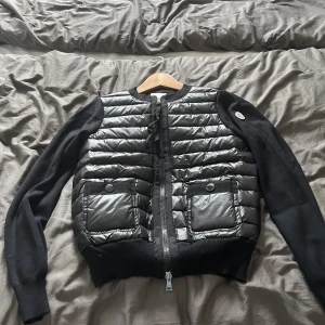 Svart dunjacka från Moncler - Hjälper min syster sälja sin svart dunjacka från Moncler med quiltad framsida och två stora fickor med knappar. Jackan har långärmade stickade ärmar och dragkedja framtill. Perfekt för dig som vill ha en stilren och exklusiv look. Använd fåtal gånger, inte säker på äkthet men allt tyder på att den är då hon köpt den sexondhand tidigare.