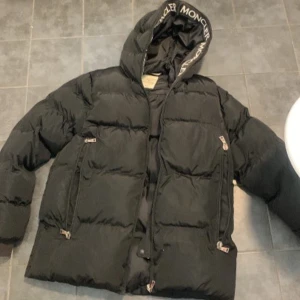 Moncler pufferjacka med huva - Säljer en svart pufferjacka från Moncler med stor huva och tydlig logga på ärmen. Jackan har dragkedjor på fickorna och insidan av huvan är dekorerad med Moncler-tryck. Perfekt för kalla dagar och riktigt snygg streetstil.