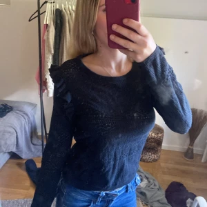 Svart stickad tröja med volangdetaljer - Snygg svart stickad tröja med volangdetaljer längs axlarna. Tröjan har rund halsringning och långa ärmar. Perfekt för dig som gillar en stilren look med en twist. Passar till både jeans och kjol.
