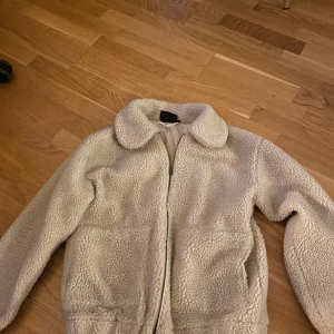 Vit teddyjacka från Tindra - Mysig vit teddyjacka från Tindra med fluffigt material och klassisk krage. Jackan har dragkedja framtill och två stora fickor. Perfekt för kalla dagar när du vill ha en chill och trendig look.