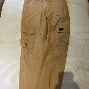 Beige cargobyxor från Vailent - Snygga beige cargobyxor från Vailent med stora fickor på sidorna och klassisk passform. Byxorna är tillverkade i bomull och har bälteshällor samt knappstängning. Perfekta för en avslappnad och trendig stil.