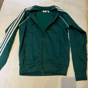 Grön Adidas hoodie med vita ränder - Snygg grön vindjacka från Adidas med klassiska vita ränder längs ärmarna och vit piping. Jackan har hel dragkedja framtill, ribbade muddar och två sidofickor. Perfekt för en sportig streetstyle-look.