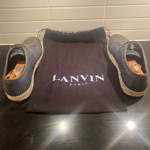 Lanvins - Snygga sneakers från Lanvin med blå och grå randig textil på sidorna och grått skinn över tån. Skorna har vita sulor och klassisk snörning. Perfekta för dig som gillar stilrena och unika detaljer i vardagslooken. Storlek UK 7. Säljs utan skoblock. Pris kan diskuteras.