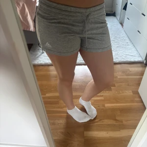Grå shorts från Adidas med snörning - Snygga grå shorts från Adidas med elastisk midja och snörning framtill. Mjukt bomullsmaterial som är skönt mot huden och passar perfekt för träning eller chill. Diskret Adidas-logga på ena benet och kort, loose passform.
