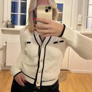 Vit kofta med svarta detaljer och guldknappar - Supersnygg vit kofta med svarta kanter och dekorativa guldknappar. Koftan har ett rutigt stickat mönster, två fejkfickor framtill och lång ärm. Perfekt för dig som gillar klassisk men trendig stil. Sitter som en S💖💖🌸🌸