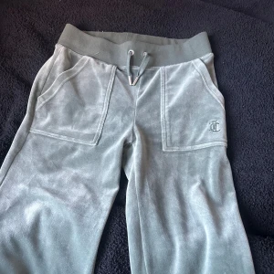 Gröna velourbyxor Juicy Couture XXS - Mysiga gröna mjukisbyxor från Juicy Couture i velour med bred resår och snörning i midjan. Stora fickor fram, bakfickor med knappar och broderad JC-logga på ena benet. Perfekta för chill dagar och snyggt avslappnad stil.