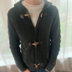 Mörkgrå stickad hoodie från Enos Jeans - Mörkgrå stickad hoodie från Enos Jeans med snygga träknappar och grovstickade detaljer längs sidorna. Tröjan har huva, långa ärmar och ribbade muddar. Perfekt för dig som gillar en chill och stilren look.
