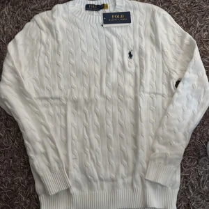 Vit kabelstickad tröja Polo Ralph Lauren - Vit kabelstickad tröja från Polo Ralph Lauren med klassisk rund halsringning och broderad marinblå logga på bröstet. Tröjan har ribbade muddar vid ärmslut och nederkant, perfekt för en clean och stilren look.