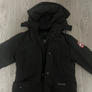Svart dunjacka från Canada Goose - Säljer en svart dunjacka från Canada Goose med huva och klassisk logga på ärmen. Jackan har flera fickor, dragkedja och knappar framtill. Yttermaterial i polyester och bomull, fodrad med vitt ankdun för extra värme. Perfekt för kalla dagar.