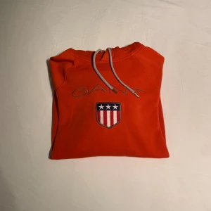 Röd hoddie från GANT - Röd hoodie från GANT med broderad logga och sköld på bröstet. Tröjan har huva med grå snören och en stor magficka. Tillverkad i mjuk bomull, perfekt för chill dagar. Klassisk och sportig look med långa ärmar.