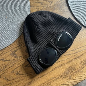 Svart ribbad mössa från C.P. Company - Säljer en svart ribbstickad mössa från C.P. Company med ikoniska svarta glasögonlinser framtill. Mössan är tillverkad i mjukt material och har en modern streetwear-look. Perfekt accessoar för att lyfta din outfit.