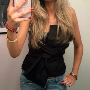 Svart omlott tubtopp med knyt - Snygg svart tubtopp med omlott och knyt i midjan. Toppen har en cool, strukturerad form och är axelbandslös vilket ger en trendig look. Perfekt att styla med jeans eller kjol för en chic outfit.