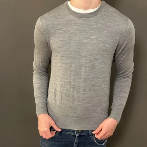 Grå stickad tröja med rund hals - Snygg grå stickad tröja med rund hals och långärmad design. Tröjan har en tunn och mjuk känsla, perfekt för lager-på-lager. Passformen är normal och den neutrala färgen gör den enkel att matcha med jeans eller andra plagg.