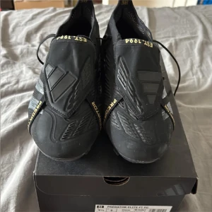 Adidas Predator Elite fotbollsskor svart/guld - Säljer ett par Adidas Predator Elite fotbollsskor i svart med guldiga detaljer och logga. Skorna har snörning, täckt plös och mönstrad ovandel. Sulan har fasta dobbar och guldtext undertill. Kommer med tillhörande Predator-påse. Perfekta för dig som vill sticka ut på planen.