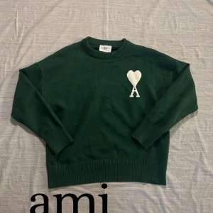 Grön stickad tröja från Ami Paris XL - Mörkgrön stickad tröja från Ami Paris i storlek XL. Tröjan har rund hals, ribbade muddar och en vit broderad logga med hjärta och bokstaven A på bröstet. Perfekt för dig som gillar stilrena och trendiga plagg.