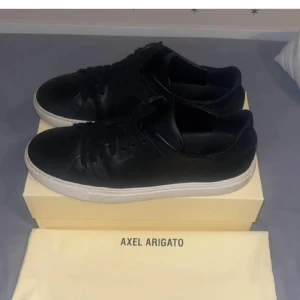 Svarta sneakers från Axel Arigato - Snygga svarta sneakers från Axel Arigato med stilren design. Skorna har svart ovandel i skinn, svarta snören och vit platt sula. Perfekta för dig som gillar minimalistisk och clean stil. Kommer med originalkartong och dustbag.