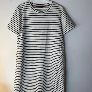 Tommy Hilfiger klänning - Vit och svart randig klänning med korta ärmar. Klänningen har en rak passform och en liten slits på sidan. En klassisk och stilren design som passar perfekt för både vardag och fest. Storlek L