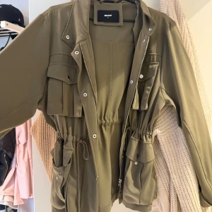 Olivgrön parkajacka från Bik Bok - Snygg olivgrön parkajacka från Bik Bok med flera fickor, snörning i midjan och dragkedja framtill. Jackan har långa ärmar och coola detaljer som tryckknappar och ficklock. Perfekt för dig som gillar streetstyle och praktiska plagg.