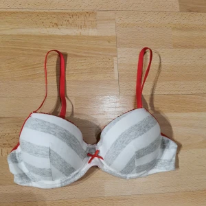 Randig Hello Kitty linnetopp 75B - Söt linnetopp från Hello Kitty med grå och vita ränder, röda axelband och en liten röd rosett framtill. Insidan är ljusrosa och har justerbara band. Perfekt för dig som gillar gulliga detaljer och färgkombinationer.