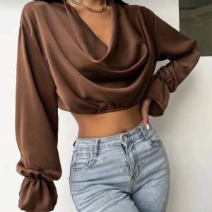 Brun croppad blus med draperad front - Säljer en brun croppad blus i satinliknande material med draperad front och v-ringning. Blusen har långa ärmar med resår vid ärmslut och en lös passform. Perfekt för dig som gillar trendiga och stilrena plagg.