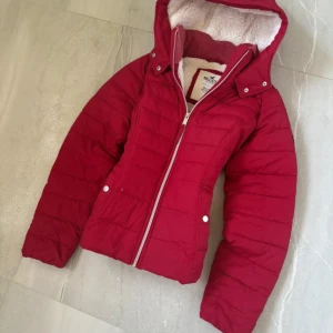 Röd pufferjacka från Hollister med huva - Riktigt snygg röd pufferjacka från Hollister med mysigt teddyfoder i huvan. Jackan har dragkedja framtill, två fickor med tryckknappar och en broderad logga på ärmen. Perfekt för kalla dagar när du vill hålla stilen.