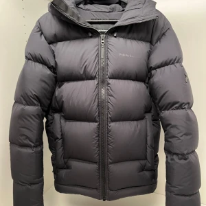 Svart dunjacka från Peak Performance - Säljer en svart dunjacka från Peak Performance med huva och dragkedja framtill. Jackan är vadderad och har en clean, sportig look med diskret logga på bröstet. Perfekt för kalla dagar och riktigt skön att bära. Passformen är normal och jackan har långa ärmar. Han på bilden är 184cm.