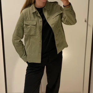 Olivgrön utilityjacka  - Snygg olivgrön utilityjacka med klassisk krage, axelklaffar och stora fickor framtill. Jackan har raka linjer, knäppning med knappar och är tillverkad i bomull. 