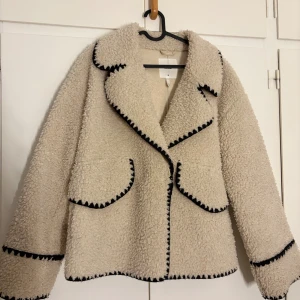 Beige teddyjacka från Object, stl 38 - Säljer en beige teddyjacka från Object i storlek 38. Jackan har svarta dekorativa sömmar längs kanterna, två fickor fram och en bred krage. Materialet är fluffigt och mjukt, perfekt för att hålla sig varm. Långärmad och loose fit för chill vibe. Använd under en vinter. 