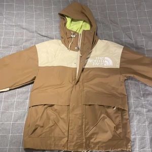 Beige och brun vindjacka The North Face - Snygg vindjacka från The North Face i beige och brunt med stor logga på bröstet. Jackan har huva med snörning, stora fickor framtill och dragkedja. Insidan är fodrad i limegrönt. Perfekt för dig som vill ha en stilren och funktionell jacka. Den är i storlek S men passar även M. Det är bara att skriva om du har några frågor.