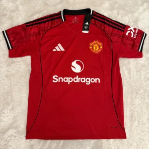 Manchester United röd matchtröja Adidas - Snygg röd Manchester United fotbollströja från Adidas med vita och svarta detaljer. Kortärmad modell med klubbmärke på bröstet och Snapdragon-logga framtill. Tillverkad i lätt och ventilerande polyester, perfekt för träning eller match.