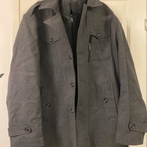 Grå ullrock med dragkedja och knappar - Helt ny. Stilren grå rock i ull med klassisk krage, axelklaffar och flera fickor framtill. Jackan har både dragkedja och knäppning, samt snygga detaljer som knappar på ärmslut och bröstficka. Perfekt för dig som vill ha en clean och tidlös look.  Den är köpt på stockhome. 