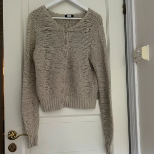 Beige stickad kofta från Bik Bok - Supermysig beige stickad kofta från Bik Bok med v-ringning och knappar framtill. Koftan har långärmade ärmar och breda ribbade muddar. Finns en fläck på ärmen.
