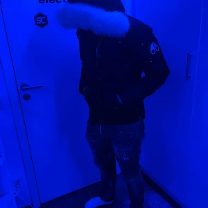 Moose Knuckles dunjacka med pälskrage - Säljer en mörk dunjacka från Moose Knuckles med stor vit pälskrage på huvan och ikonisk logga på ärmen. Jackan har dragkedja, knappar och flera fickor. Perfekt för kalla dagar och riktigt snygg streetstil.