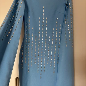 Ljusblå långklänning med paljettdetaljer - Ljusblå långklänning med långa ärmar och glittrande paljettdetaljer i vertikala rader framtill och på ärmarna. Klänningen har en elegant, rak passform och är tillverkad i ett mjukt, slätt tyg. Dragkedja i ryggen för enkel påklädning.