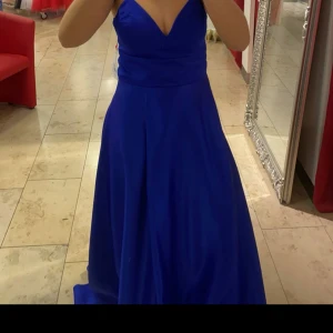 mörkblå  långklänning med snörning - Elegant mörkblå långklänning i glansigt material med smala axelband, v-ringad urringning och snygg snörning i ryggen. Klänningen har en figurnära överdel och ett svepande, utsvängt kjolparti som ger en lyxig känsla. Perfekt för bal eller speciella tillfällen. Använd en gång och ser ut som ny.