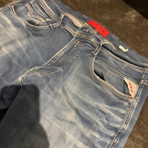 Replay Anbass ljusblå jeans - Säljer ett par Replay Anbass jeans i ljusblå tvätt med snygga slitningar och klassiska detaljer. Jeansen har fem fickor, Replay-logga på myntfickan och bakficka samt röd etikett i midjan. Skön passform och perfekt för dig som gillar en avslappnad stil.